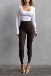 A female model wearing a white long-sleeve crop top and dark brown full-length leggings.