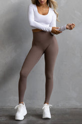 A woman standing against a grey background, wearing full-length leggings in a soft umber color with a white long-sleeve top.