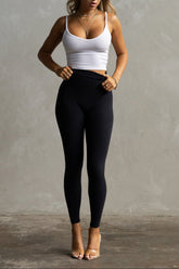 A woman wearing a white tank top and black full-length leggings, standing against a gray background.