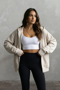 Woman wearing a beige hoodie with thumbholes, white tank, and black leggings against a gray background