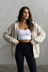 Woman wearing a beige hoodie with thumbholes, white tank, and black leggings against a gray background