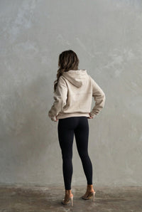 Person wearing a beige hoodie and black leggings standing against a plain wall.