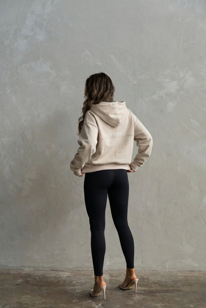 Person wearing a beige hoodie and black leggings standing against a plain wall.