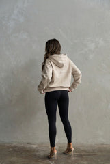 Person wearing a beige hoodie and black leggings standing against a plain wall.