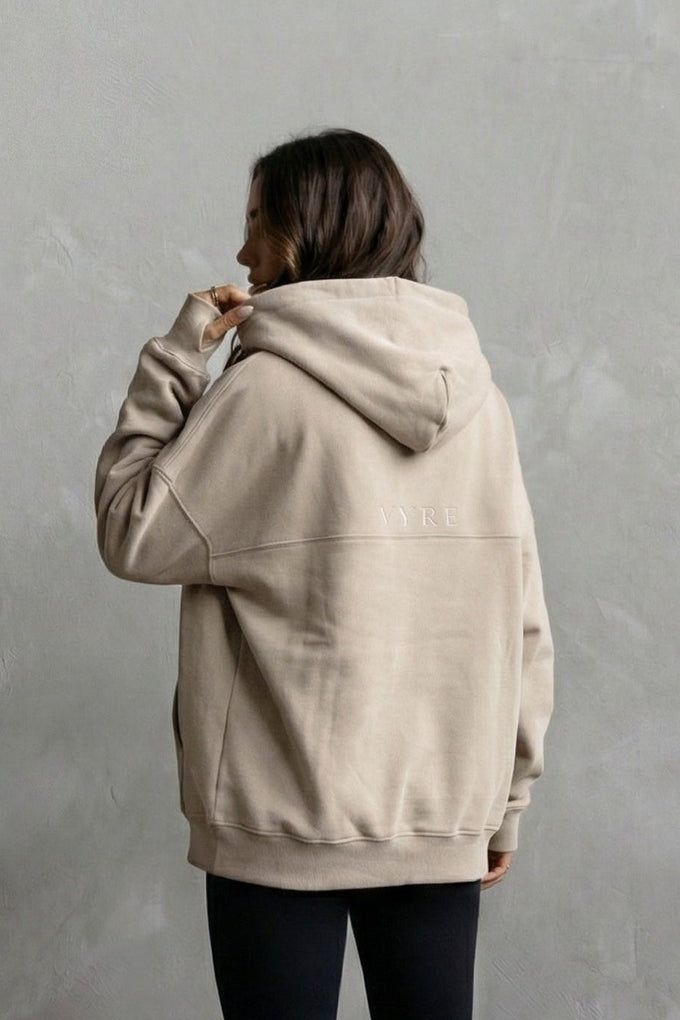 Person wearing a beige hoodie and black leggings against a gray background