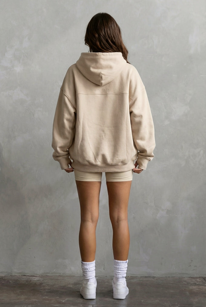 Person wearing a beige hoodie and shorts set against a gray concrete wall.