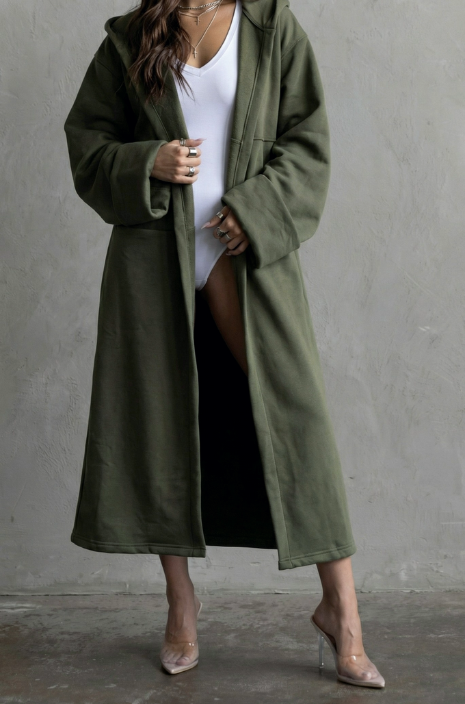 Woman wearing a long olive green coat over a white top and black skirt on a gray background